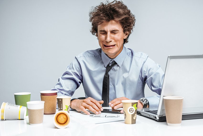 A man crying by his laptop with several empty coffee cups on his desk.